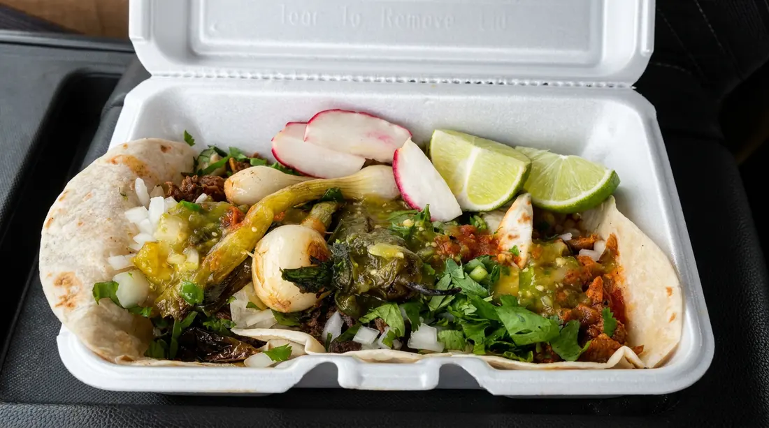 An open takeout box filled with tacos, lime, radish, and onion